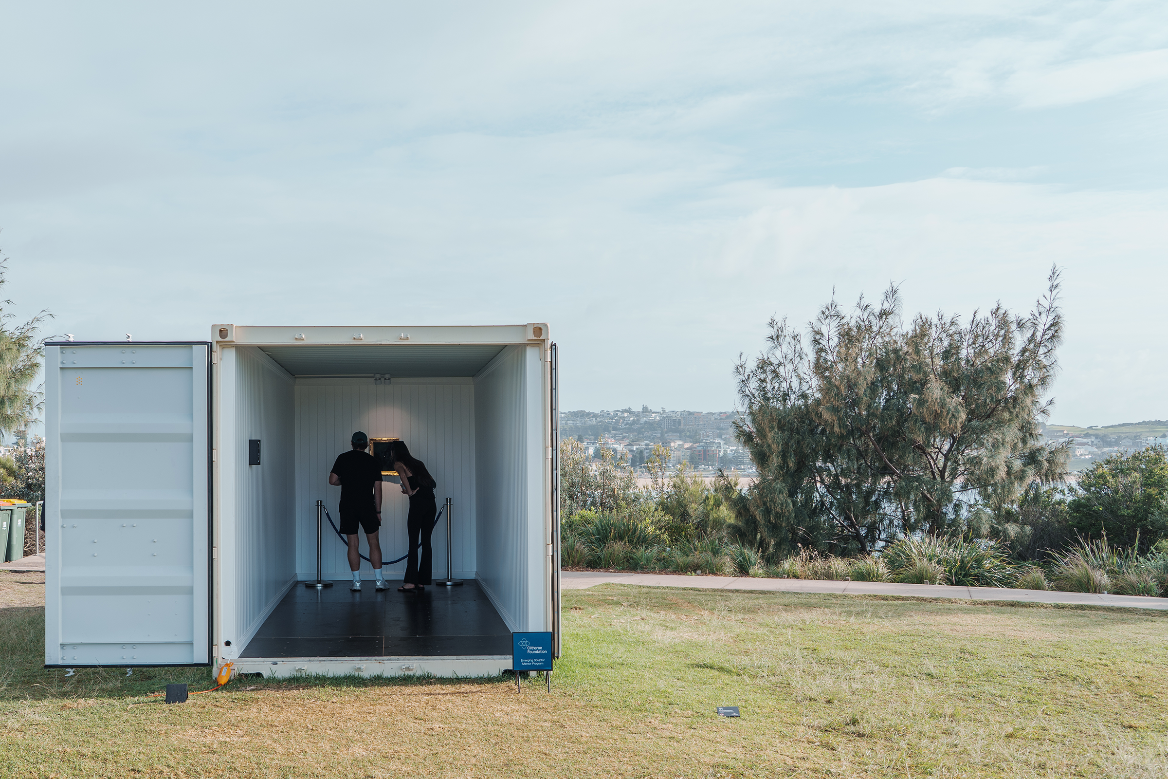 Two people looking at the display inside the container, with the surrounding park and sky visible.